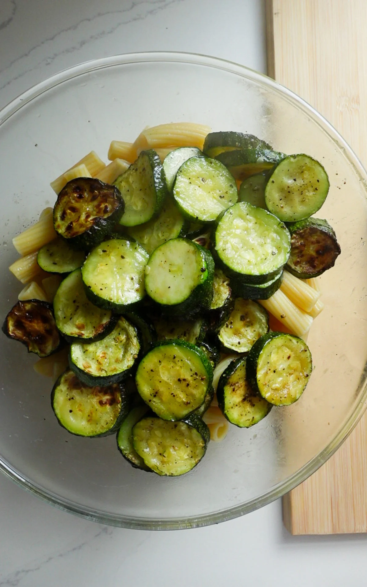A glass bowl contains an easy zucchini pasta salad with penne and sliced, roasted zucchini. The lightly browned, seasoned veggies are topped with a drizzle of Parmesan Dressing. The bowl rests on a white surface near a wooden cutting board.