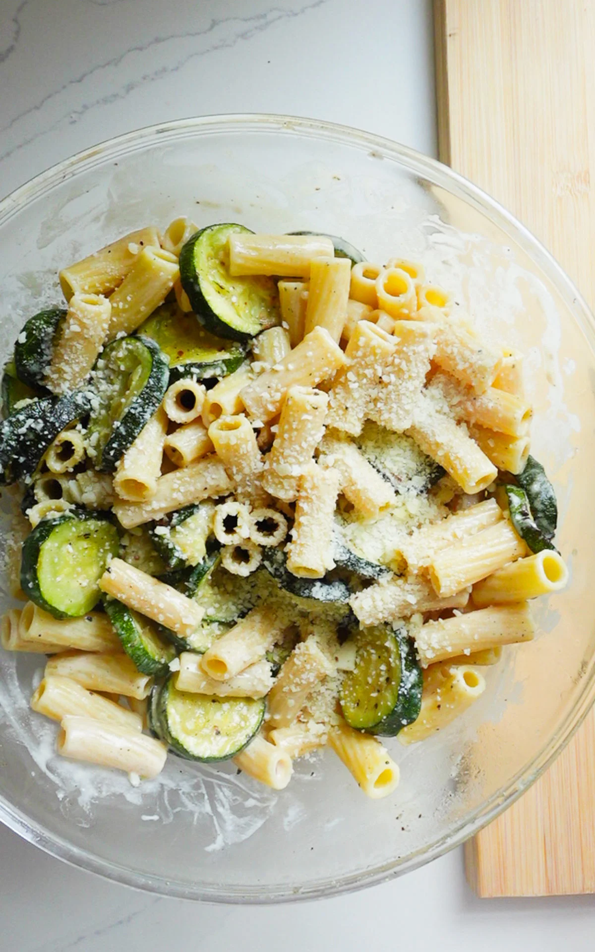 A clear glass bowl filled with fresh zucchini pasta salad, rigatoni, and sprinkled grated Parmesan cheese sits on a white marble countertop next to a wooden cutting board.