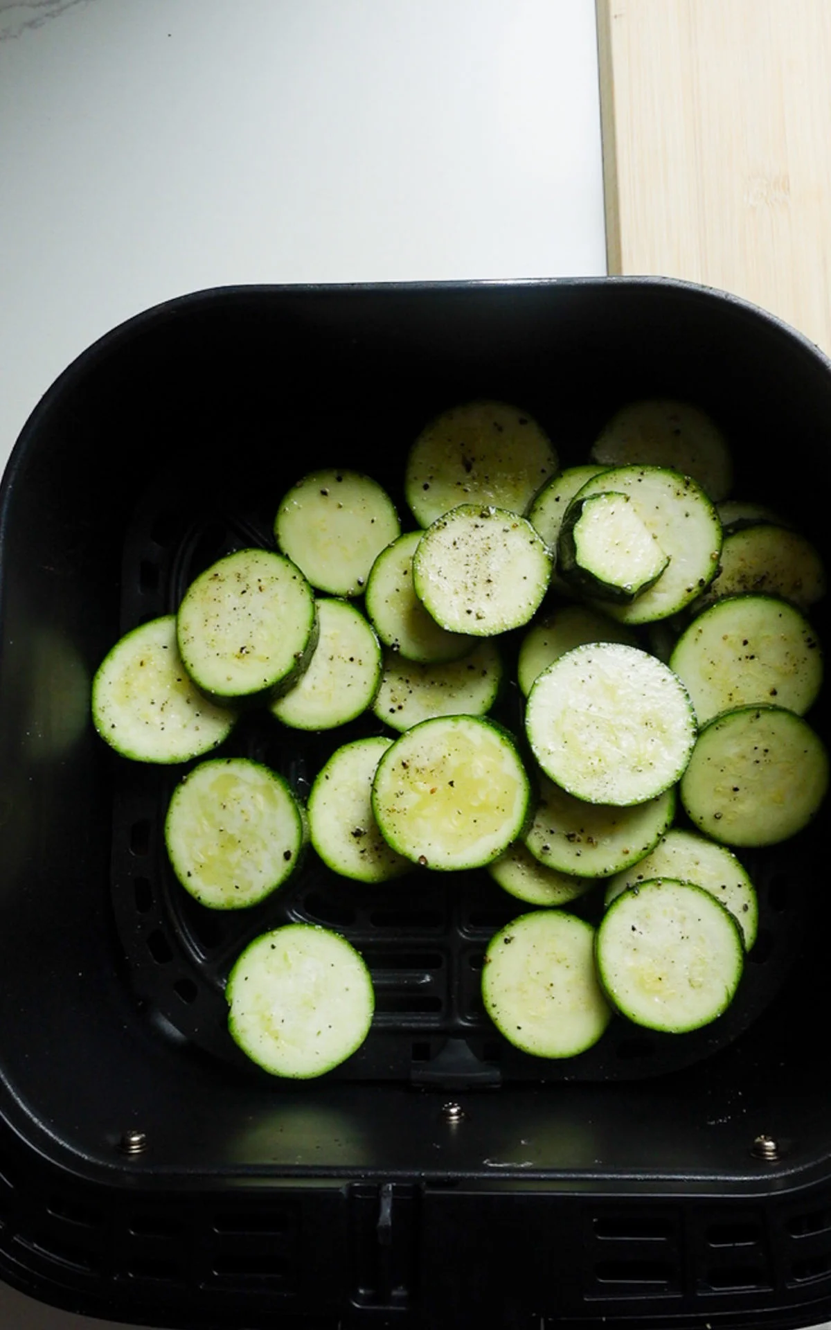 Sliced zucchini rounds, seasoned with pepper and possibly salt, are arranged in a black air fryer basket for cooking—perfect for tossing into a fresh Zucchini Pasta Salad or drizzling with Parmesan Dressing. A wooden cutting board sits partially visible on the right.