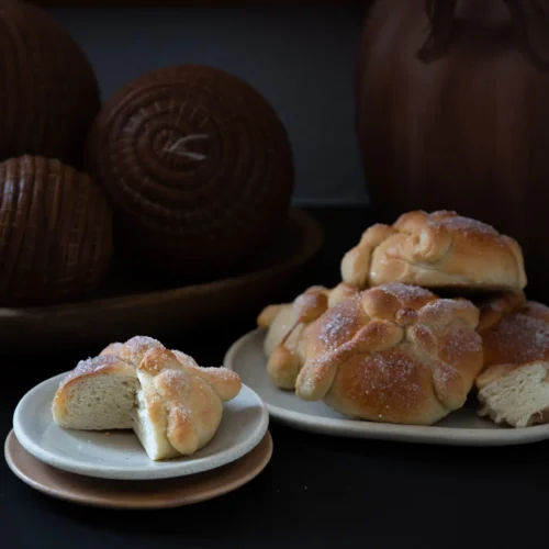 A small plate holds a halved sweet bread roll dusted with sugar, showing its soft interior. Next to it, a larger plate displays more sugared rolls. In the background are brown decorative spheres and clay pottery, all set against a dark surface.