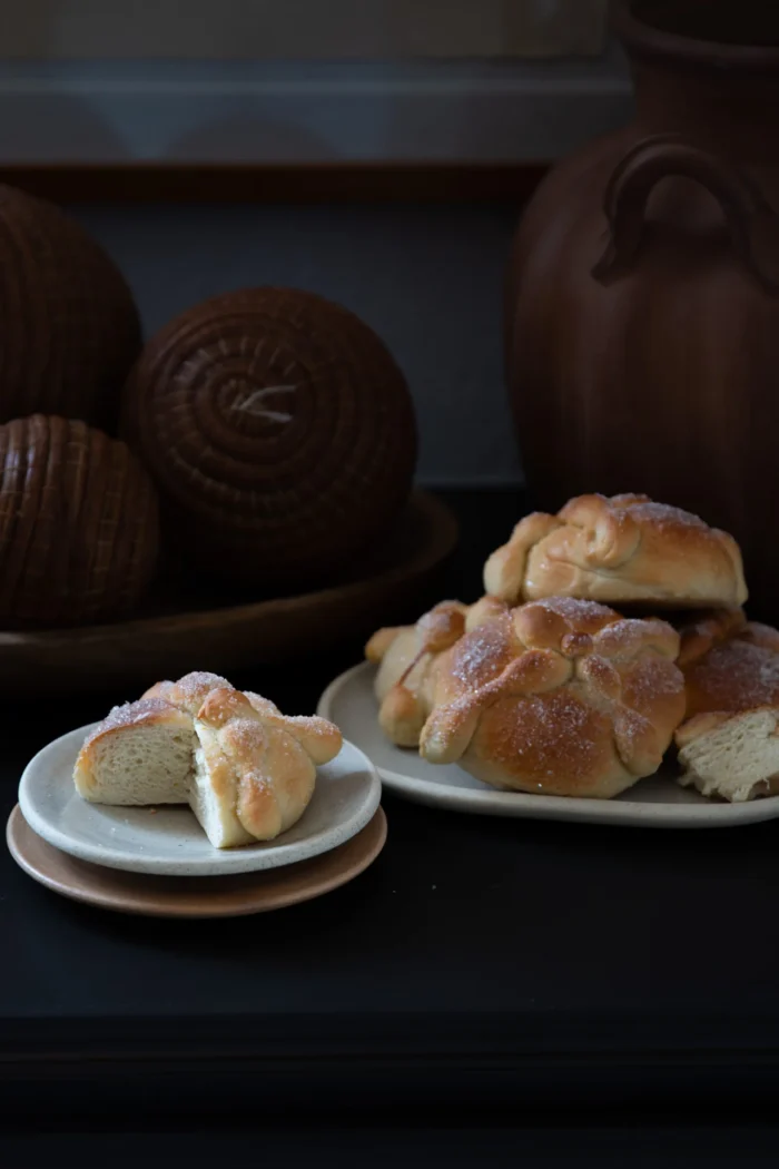 A small plate holds a halved sweet bread roll dusted with sugar, showing its soft interior. Next to it, a larger plate displays more sugared rolls. In the background are brown decorative spheres and clay pottery, all set against a dark surface.