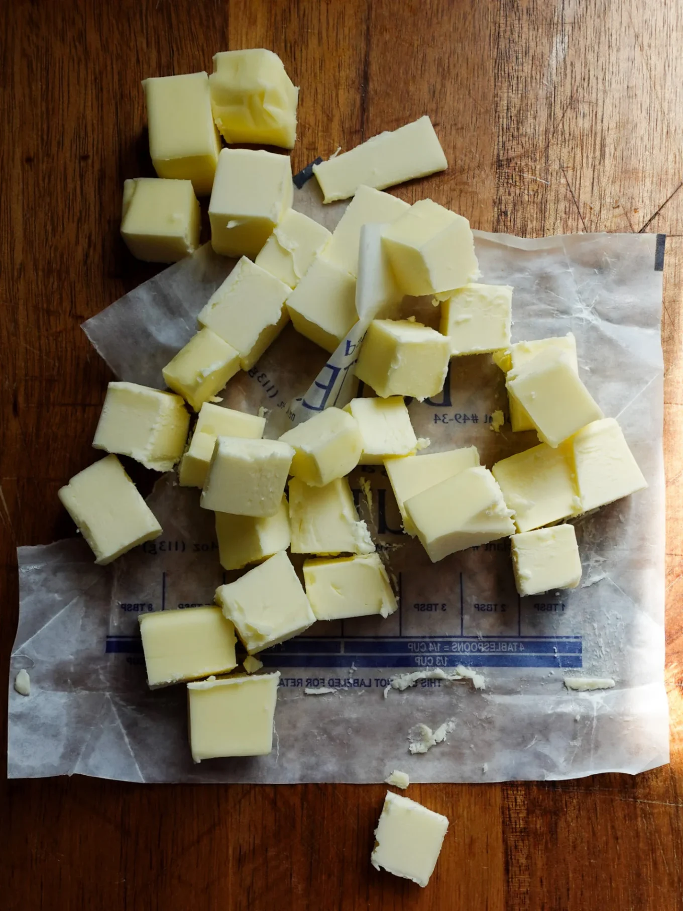 Bright yellow cubes of butter are scattered on a butter wrapper atop a wooden surface. Perfect for preparing authentic pan de muerto, the cubes rest on faint blue measurement markings, while natural light casts soft shadows.