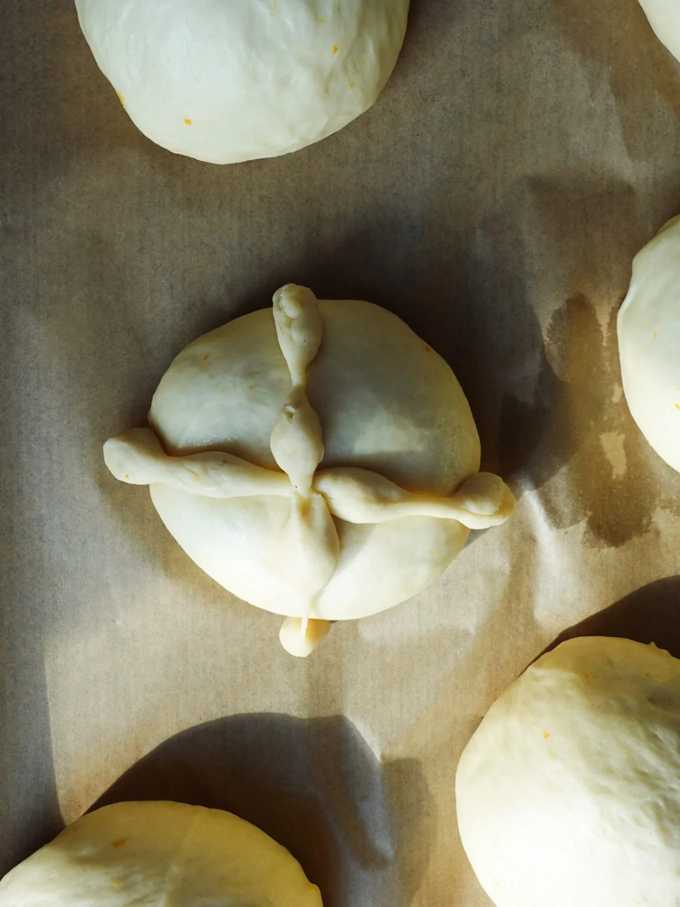 Round dough balls on brown parchment paper, with one centered bun decorated with two twisted dough strips forming a cross shape on top—an authentic pan de muerto, the traditional Mexican bread for Day of the Dead, highlighted in soft light.