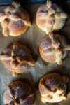 Six round pieces of authentic pan de muerto, golden brown and dusted with sugar, sit on parchment paper. Each traditional Mexican bread features bone-shaped dough decorations on top, arranged in a crossed pattern.