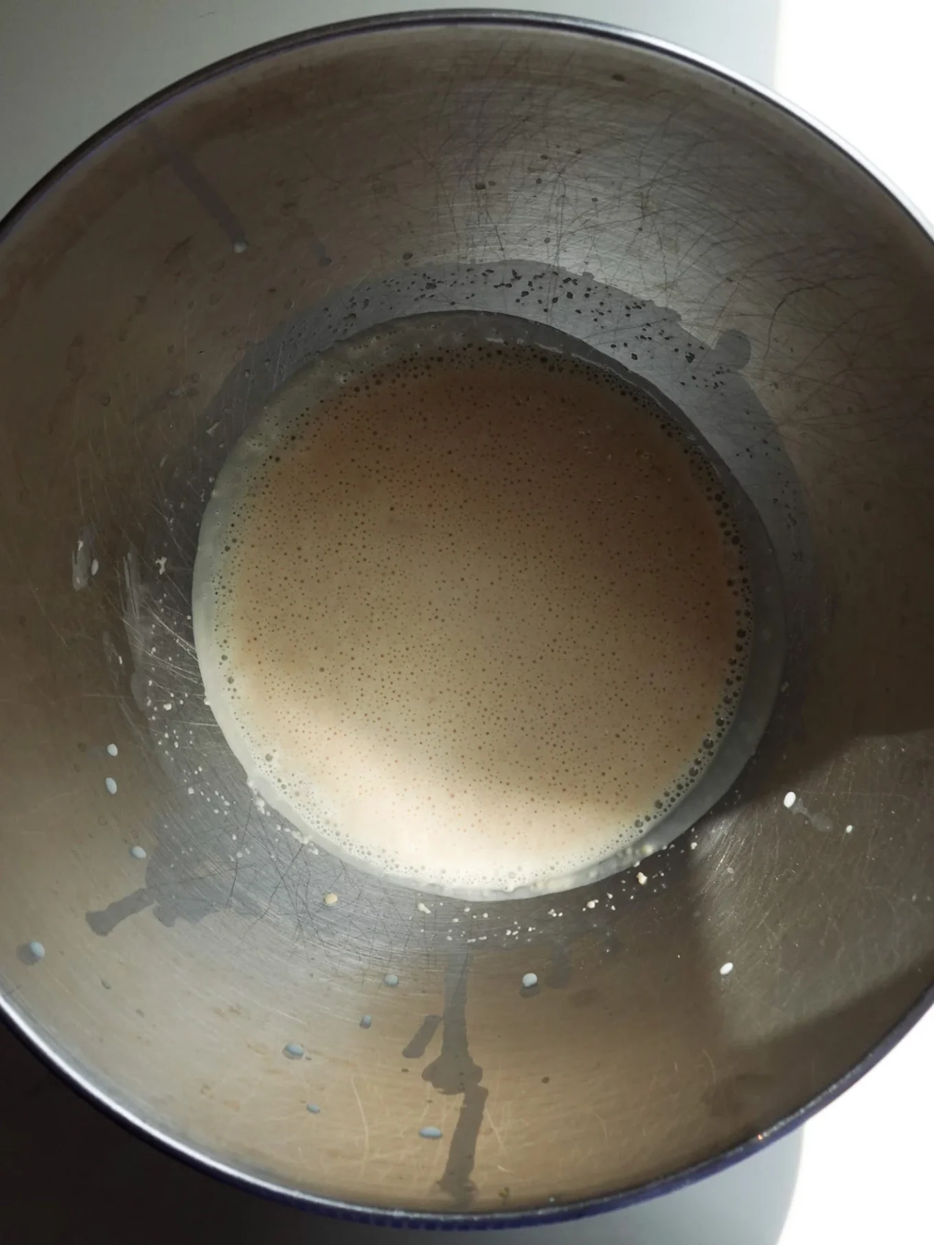A metal mixing bowl containing a light tan, frothy authentic pan de muerto batter. Sunlight highlights half of the bowl, casting shadows and revealing splatter marks and scratches on the interior sides. The bowl sits on a white surface.