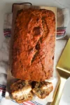 A golden-brown loaf of moist banana bread with a cracked top sits on a wooden cutting board. Two slices are cut in front, showing pieces of walnut inside. The setup rests on a textured towel with red, white, and black stripes beside a gold cake server.
