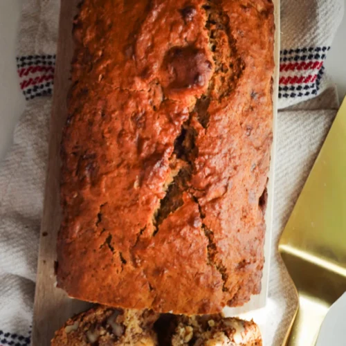A golden-brown loaf of moist banana bread with a cracked top sits on a wooden cutting board. Two slices are cut in front, showing pieces of walnut inside. The setup rests on a textured towel with red, white, and black stripes beside a gold cake server.