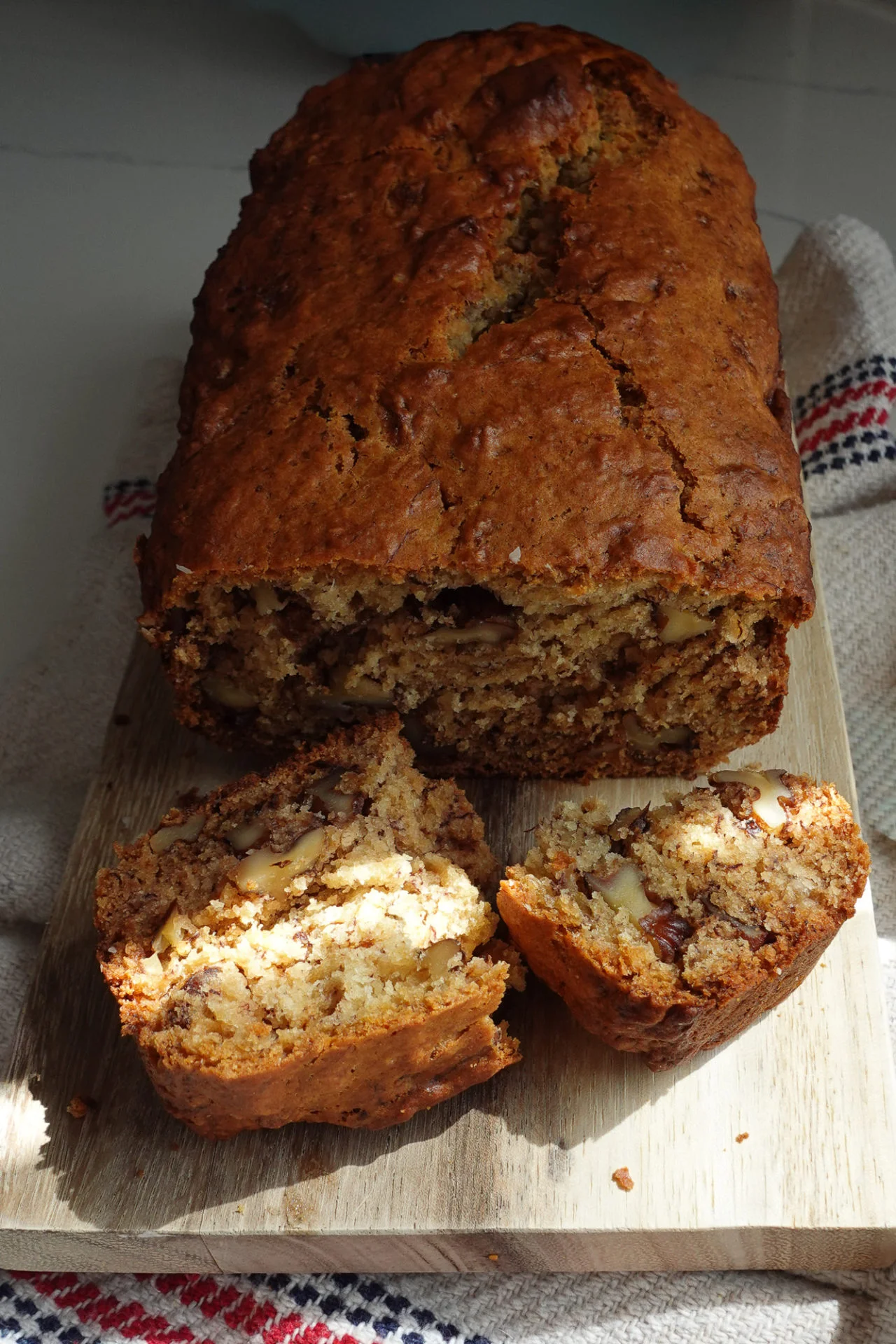 A loaf of Walnut Banana Bread with chopped walnuts sits on a wooden board. Two slices are cut in front, revealing a moist, nutty interior. The bread is golden-brown, resting on a textured kitchen towel with red and blue stripes.