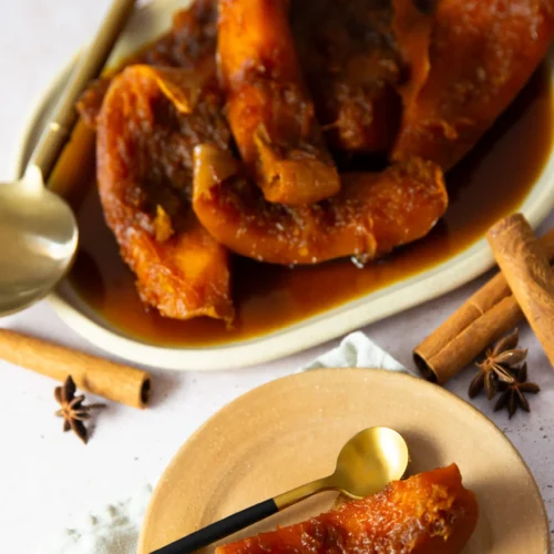 Slices of candied pumpkin, or Calabaza en Tacha, are arranged on an oval plate, with one piece on a round tan plate. A gold spoon rests nearby, while cinnamon sticks and star anise hint at the warm spices in this classic Mexican dessert.