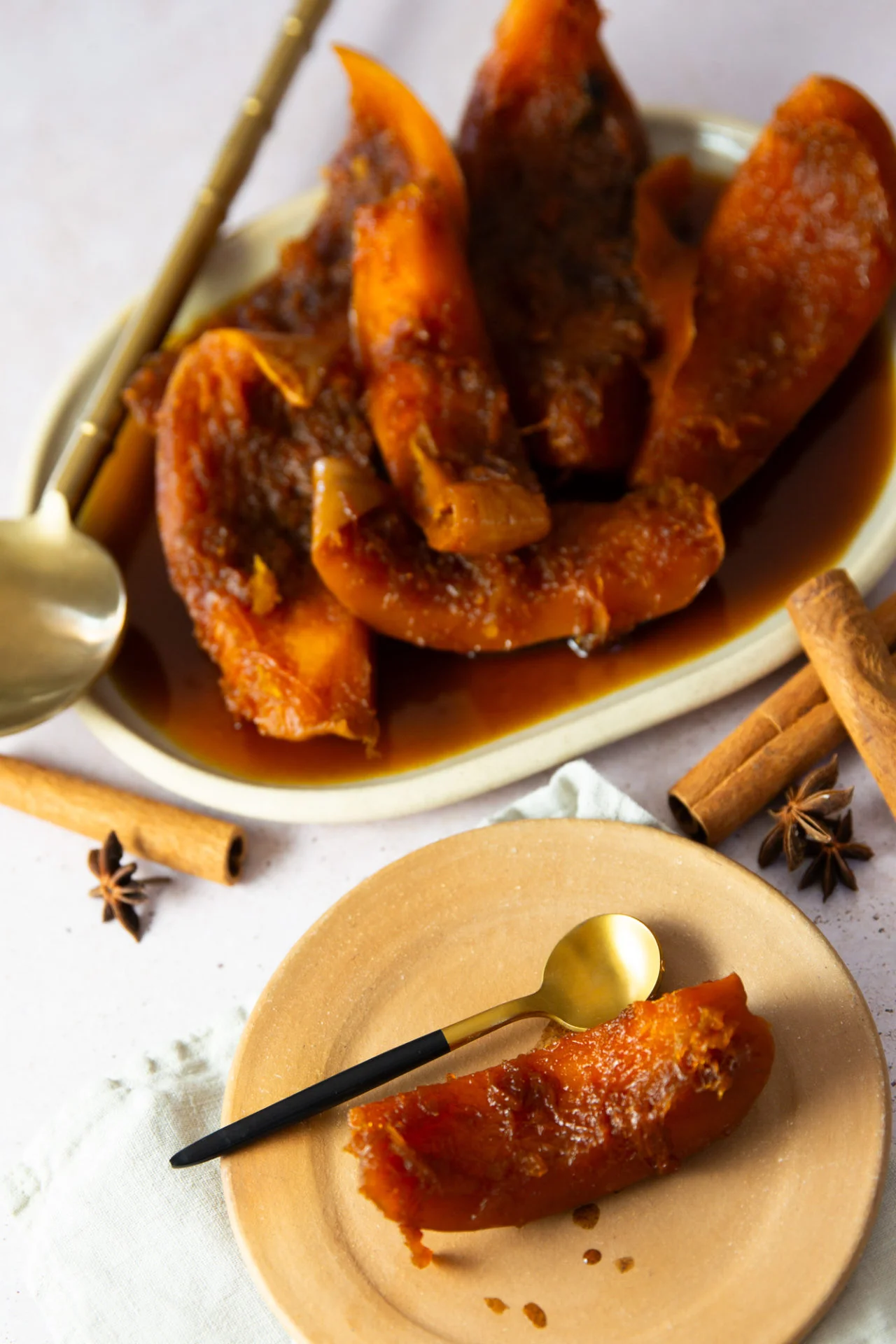 Slices of candied pumpkin, or Calabaza en Tacha, are arranged on an oval plate, with one piece on a round tan plate. A gold spoon rests nearby, while cinnamon sticks and star anise hint at the warm spices in this classic Mexican dessert.
