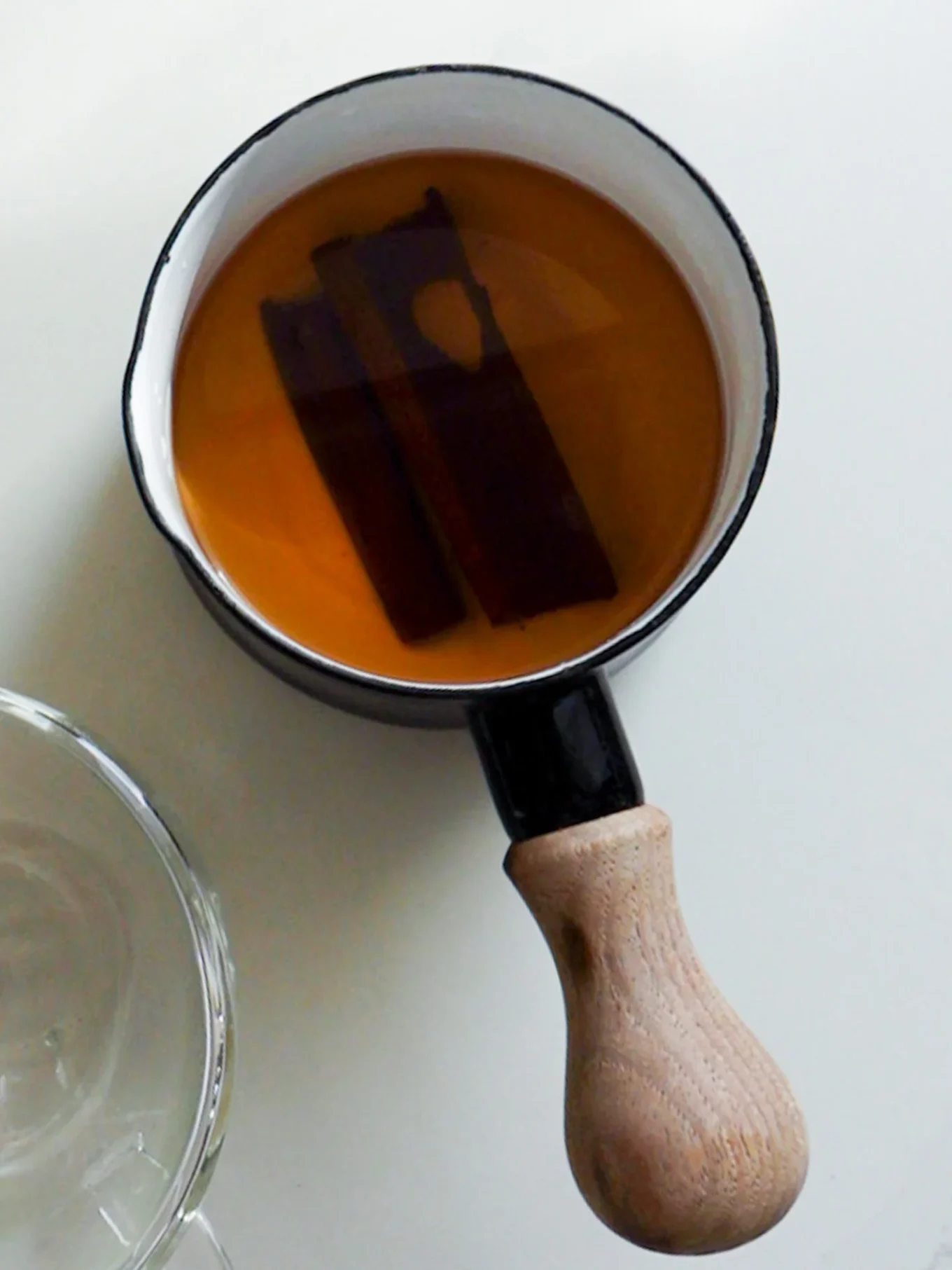 A small white saucepan with a wooden handle contains amber-colored Mexican Cinnamon Tea, or Té de Canela, with two cinnamon sticks. The saucepan sits on a white surface, and part of a clear glass bowl is visible to the left in soft, natural lighting.
