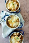 Tres pequeños cuencos con asas azules de sopa de cebolla vegana, cubiertos de queso fundido y pan, reposan sobre una servilleta blanca con dos cucharas de plata a su lado sobre una superficie de mármol claro. Una ramita de tomillo adorna los cuencos.