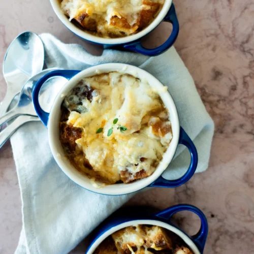 Tres pequeños cuencos con asas azules de sopa de cebolla vegana, cubiertos de queso fundido y pan, reposan sobre una servilleta blanca con dos cucharas de plata a su lado sobre una superficie de mármol claro. Una ramita de tomillo adorna los cuencos.