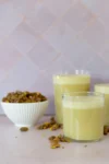 Three glasses of creamy, pale green pistachio milk are placed on a light surface. A small white bowl filled with shelled pistachios sits nearby, with some nuts scattered around. The background consists of light purple tiles.