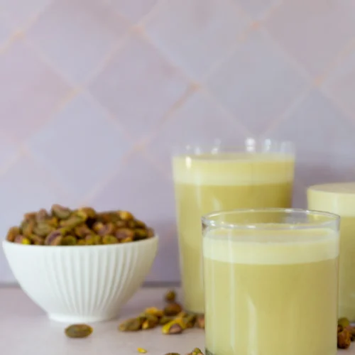Three glasses of creamy, pale green pistachio milk are placed on a light surface. A small white bowl filled with shelled pistachios sits nearby, with some nuts scattered around. The background consists of light purple tiles.