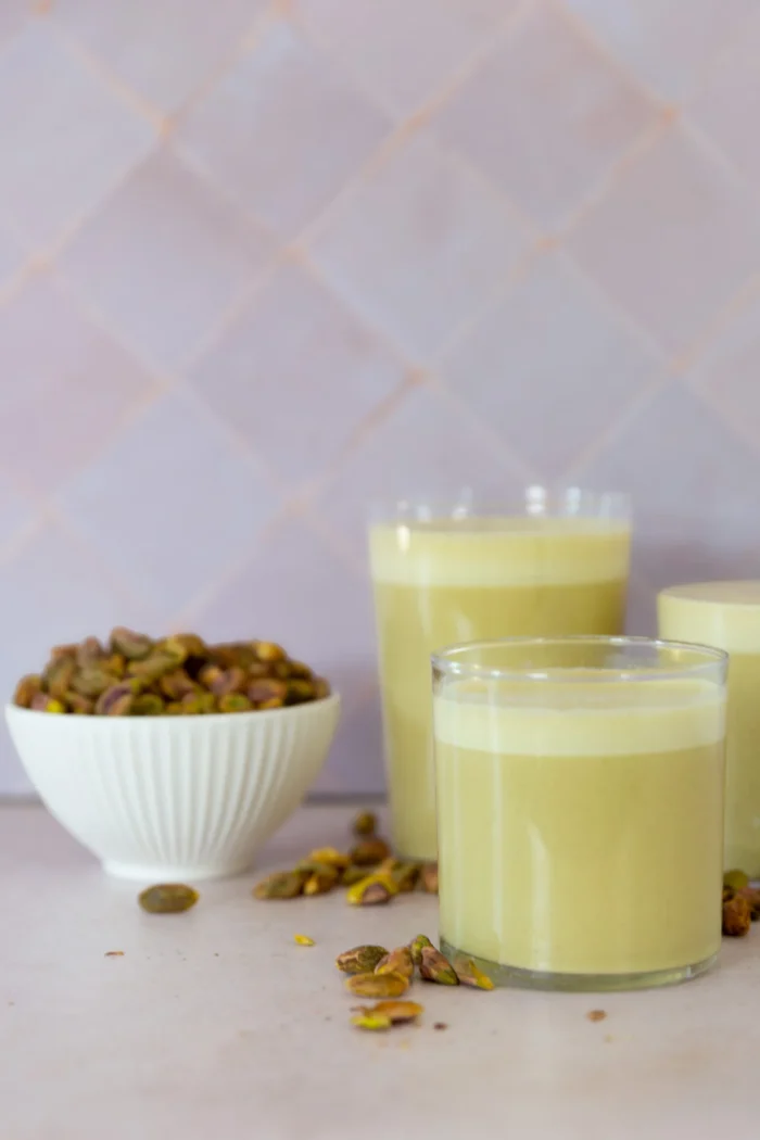 Three glasses of creamy, pale green pistachio milk are placed on a light surface. A small white bowl filled with shelled pistachios sits nearby, with some nuts scattered around. The background consists of light purple tiles.