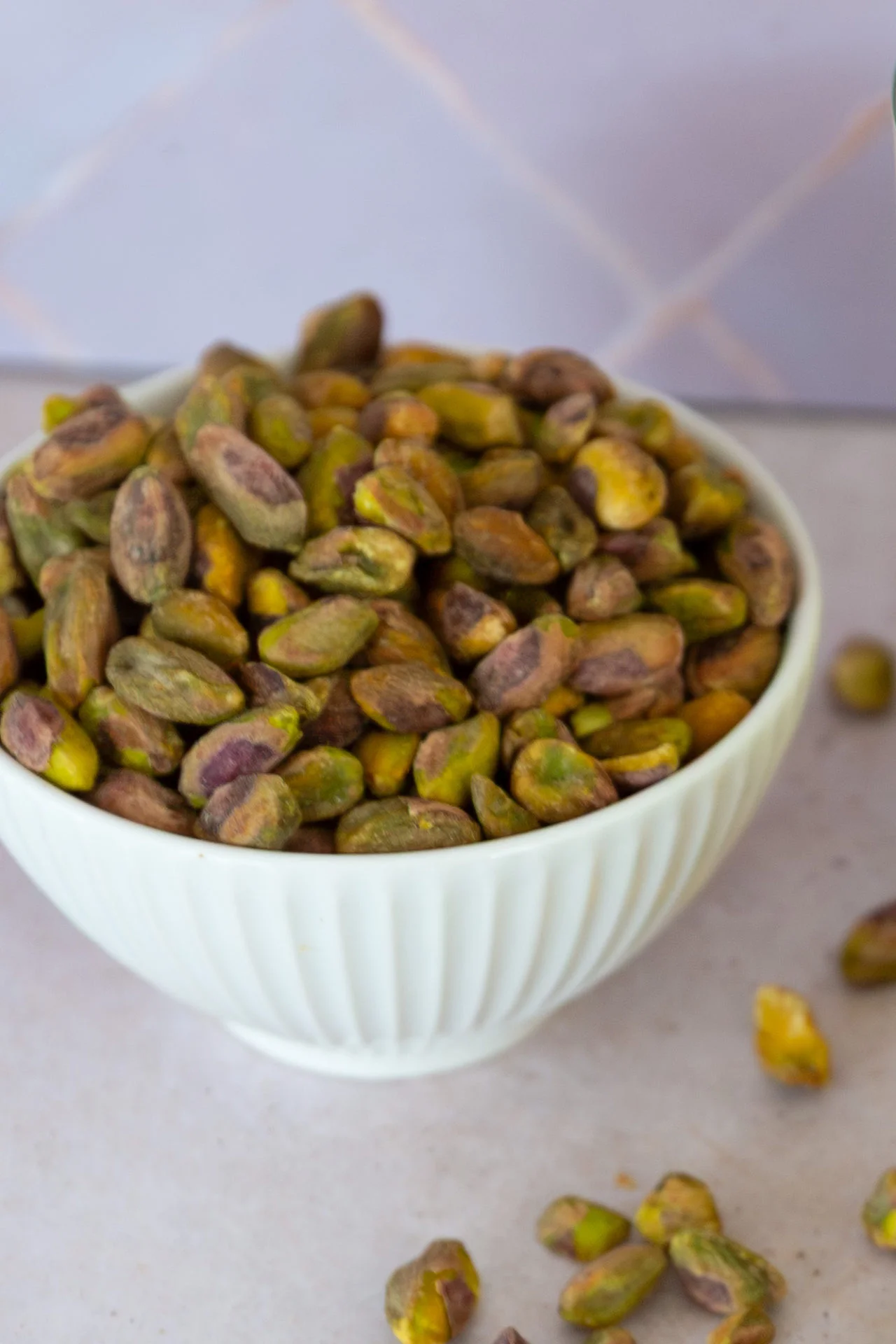 A white ceramic bowl filled with unshelled pistachios sits on a light surface, with a few scattered nearby—perfect for making homemade pistachio milk. The background features a soft, purple tile pattern, highlighting the naturally creamy green and purple-brown hues.