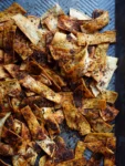 A close-up of crispy Spicy Tortilla Chips scattered on a metal baking sheet. Baked in the oven, the strips are seasoned with a dark, speckled spice mix, giving them a rich, flavorful appearance as some curl and overlap each other.
