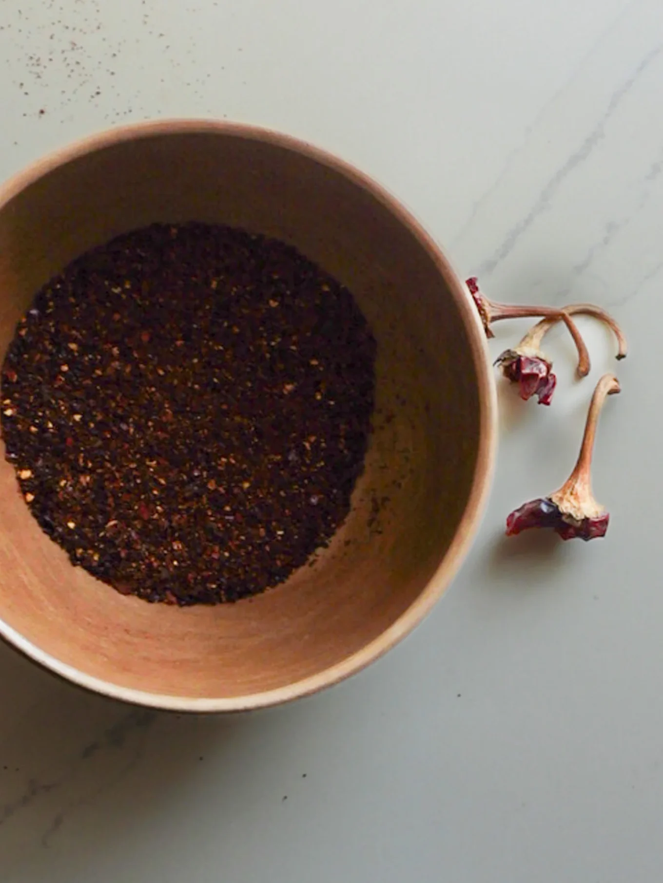 A wooden bowl filled with finely ground, dark red chili powder sits on a light-colored surface. To the right of the bowl are three dried chili stems—perfect for adding heat to oven-baked Spicy Tortilla Chips. Some powder is scattered near the rim.