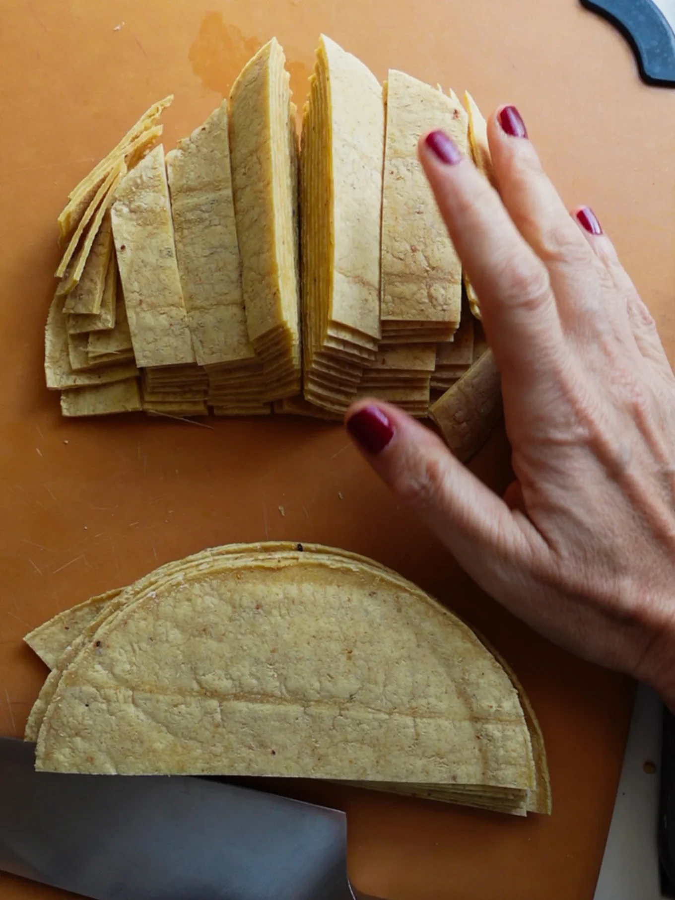 A hand with red nail polish steadies a stack of corn tortillas being sliced into strips on a brown cutting board—perfect for making Spicy Tortilla Chips in the oven. A knife rests nearby, with neatly piled tortilla strips ready to bake.