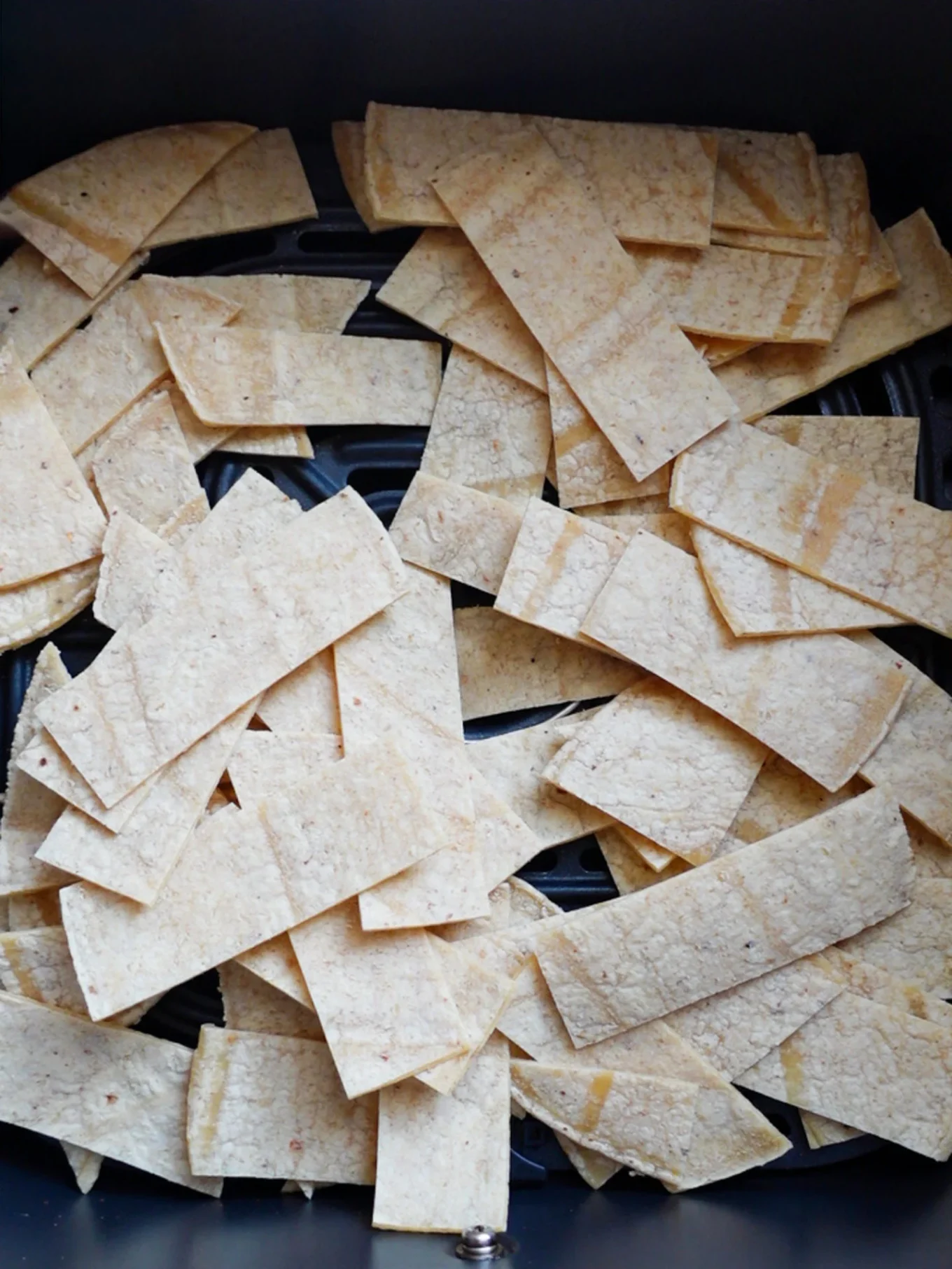 Rectangular pieces of uncooked corn tortilla are scattered in a single layer inside an air fryer basket, ready to become crispy Spicy Tortilla Chips. The pale, rough-textured tortillas have slightly uneven edges and overlap in some spots.