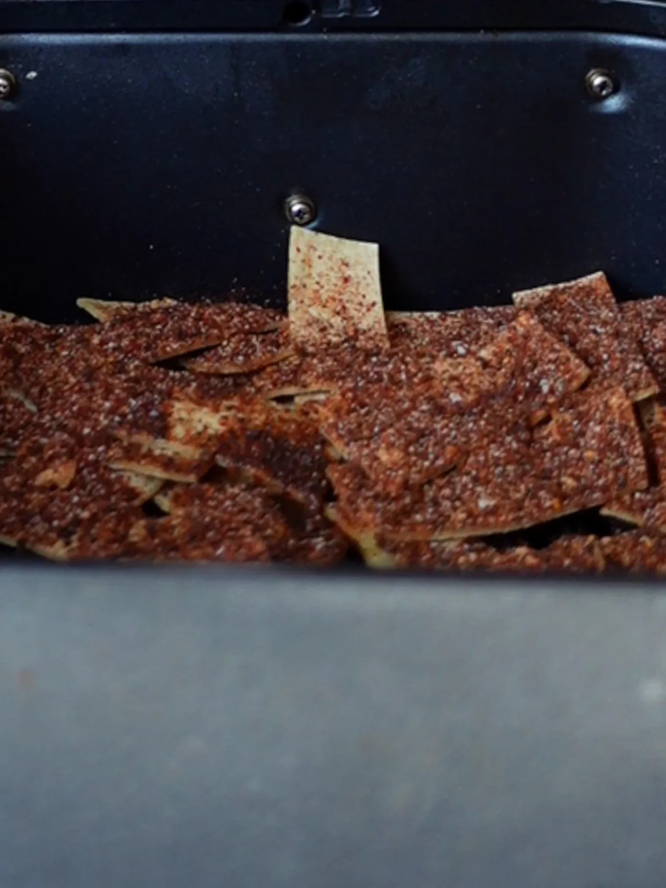 Rectangular spicy tortilla chips, coated with a visible layer of dark seasoning like chili powder and salt, are arranged inside a black air fryer. The golden-brown crackers showcase a bold spice blend for extra flavor.