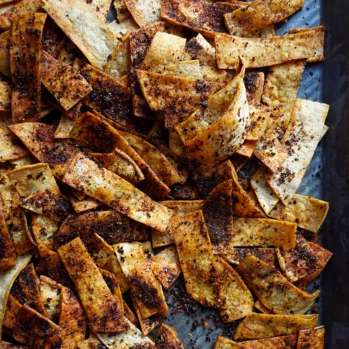 A close-up of crispy Spicy Tortilla Chips scattered on a metal baking sheet. Baked in the oven, the strips are seasoned with a dark, speckled spice mix, giving them a rich, flavorful appearance as some curl and overlap each other.