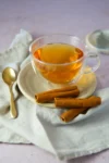 A clear glass cup of amber-colored Mexican Cinnamon Tea (Té de Canela) sits on a beige plate, with three cinnamon sticks beside it. A gold spoon rests nearby on a light cloth napkin, and a matching saucer is partially visible in the background.