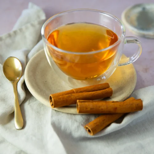 A clear glass cup of amber-colored Mexican Cinnamon Tea (Té de Canela) sits on a beige plate, with three cinnamon sticks beside it. A gold spoon rests nearby on a light cloth napkin, and a matching saucer is partially visible in the background.