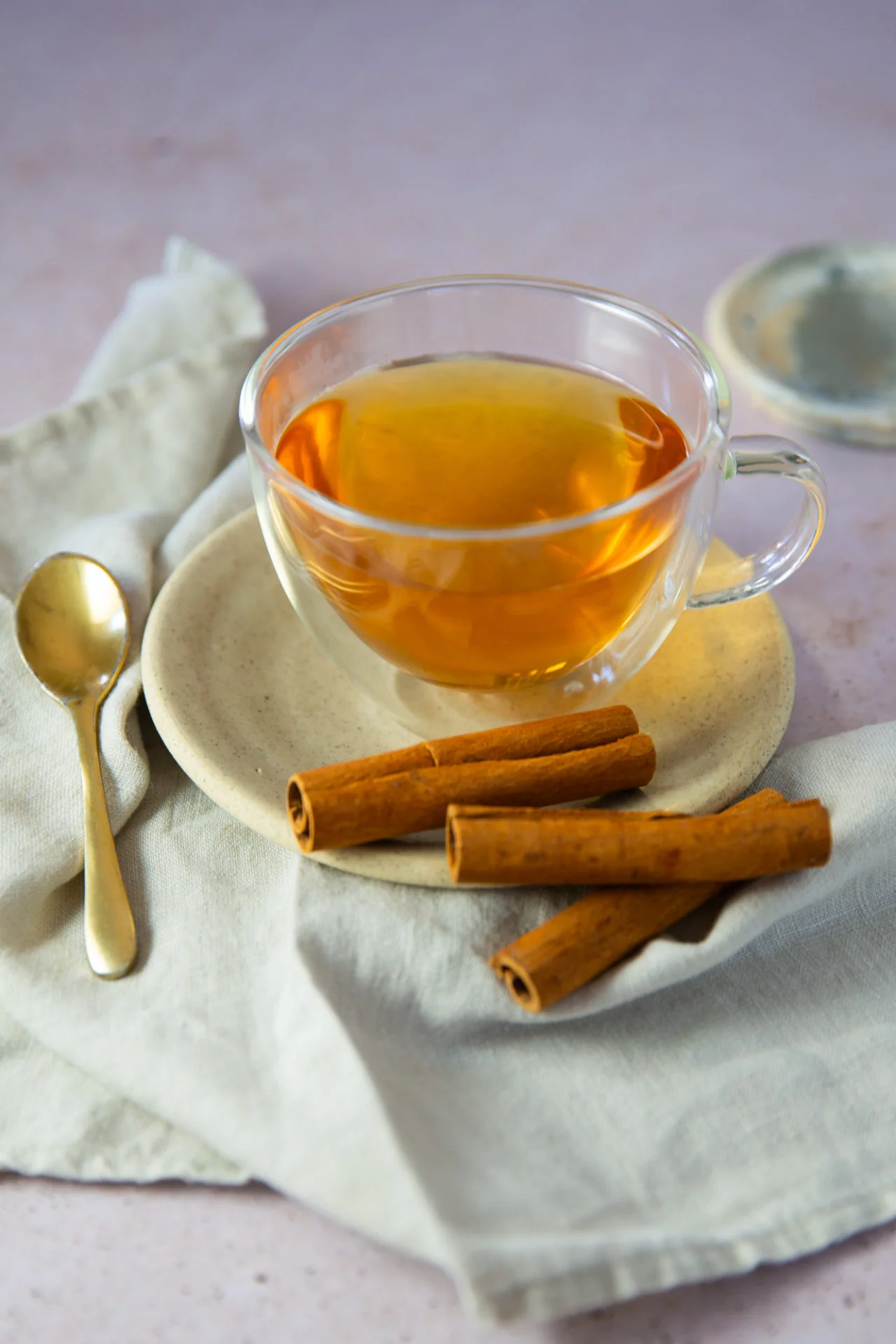 A clear glass cup of amber-colored Mexican Cinnamon Tea (Té de Canela) sits on a beige plate, with three cinnamon sticks beside it. A gold spoon rests nearby on a light cloth napkin, and a matching saucer is partially visible in the background.