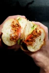 Two hands hold a sandwich sliced in half, revealing seasoned Mexican chorizo, sliced tomatoes, red onion, green lettuce, and mayonnaise inside a soft, round white bun. The dark background brings the torta-style sandwich into sharp focus.