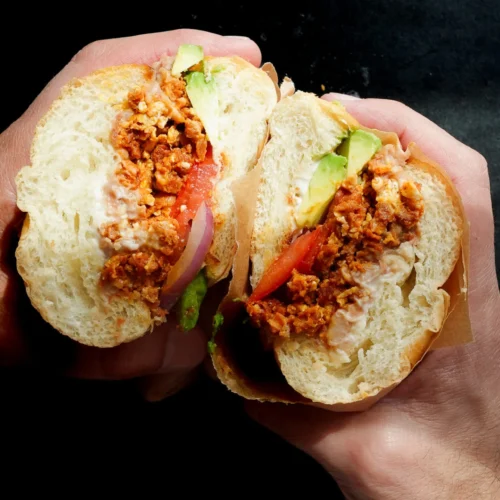 Two hands hold a sandwich sliced in half, revealing seasoned Mexican chorizo, sliced tomatoes, red onion, green lettuce, and mayonnaise inside a soft, round white bun. The dark background brings the torta-style sandwich into sharp focus.