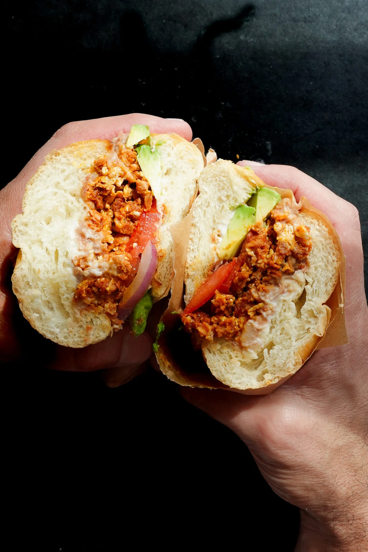 Two hands hold a sandwich sliced in half, revealing seasoned Mexican chorizo, sliced tomatoes, red onion, green lettuce, and mayonnaise inside a soft, round white bun. The dark background brings the torta-style sandwich into sharp focus.