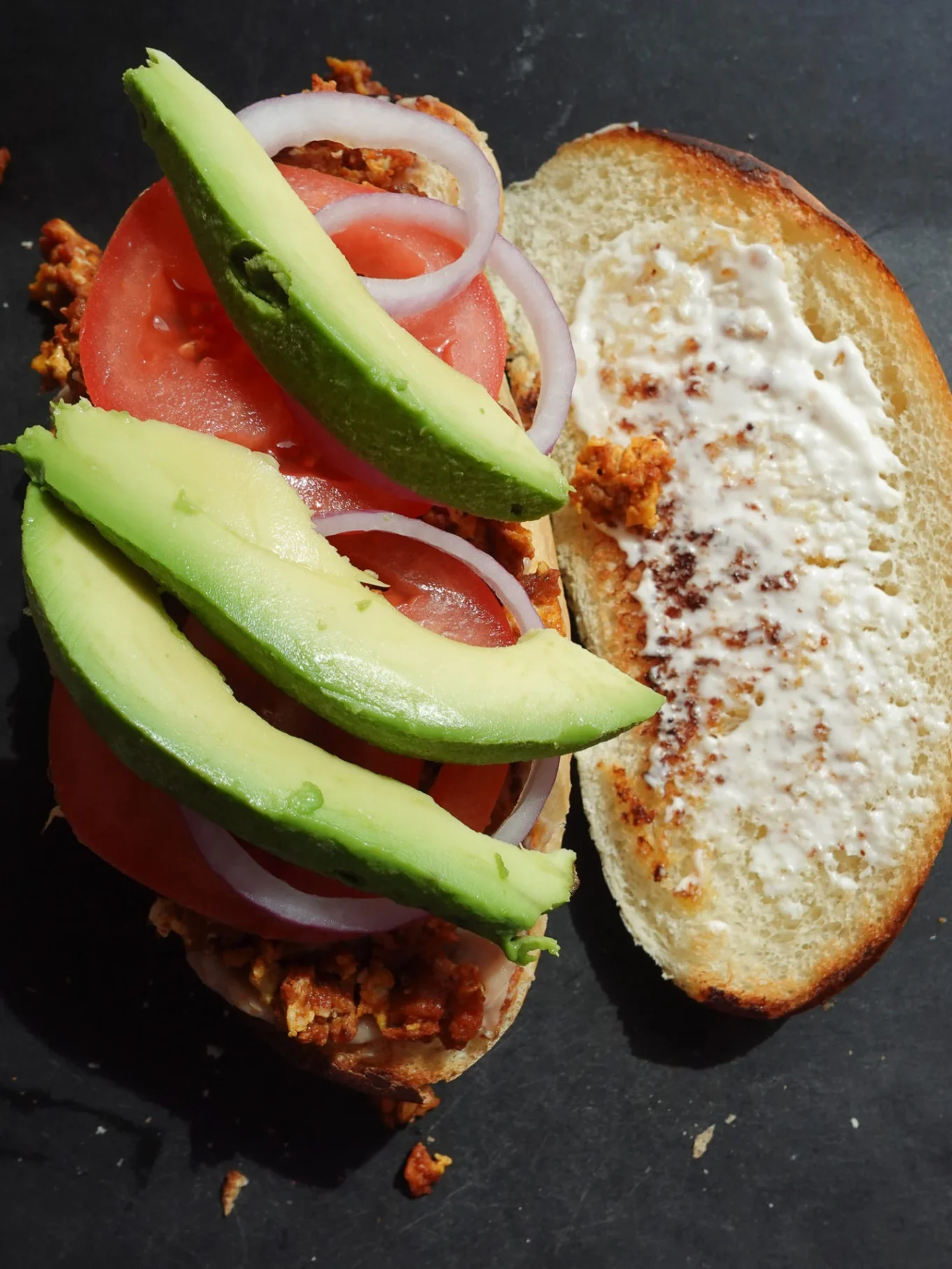 An open-faced Mexican sandwich on dark bread features cooked crumbled chorizo, tomato slices, raw onion rings, and avocado on one half, while the other is spread with mayonnaise. This Torta de Chorizo rests on a dark surface, lit from above.
