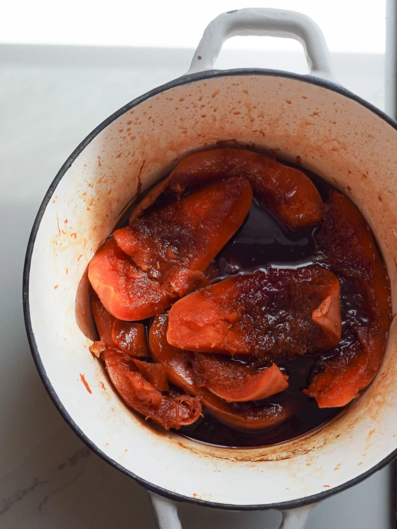 A white enameled pot on a light countertop contains sweet pumpkin, resembling Calabaza en Tacha, in a dark, seasoned liquid. The tender pieces tint the pot’s interior, echoing this classic Mexican dessert’s rich colors and aroma.