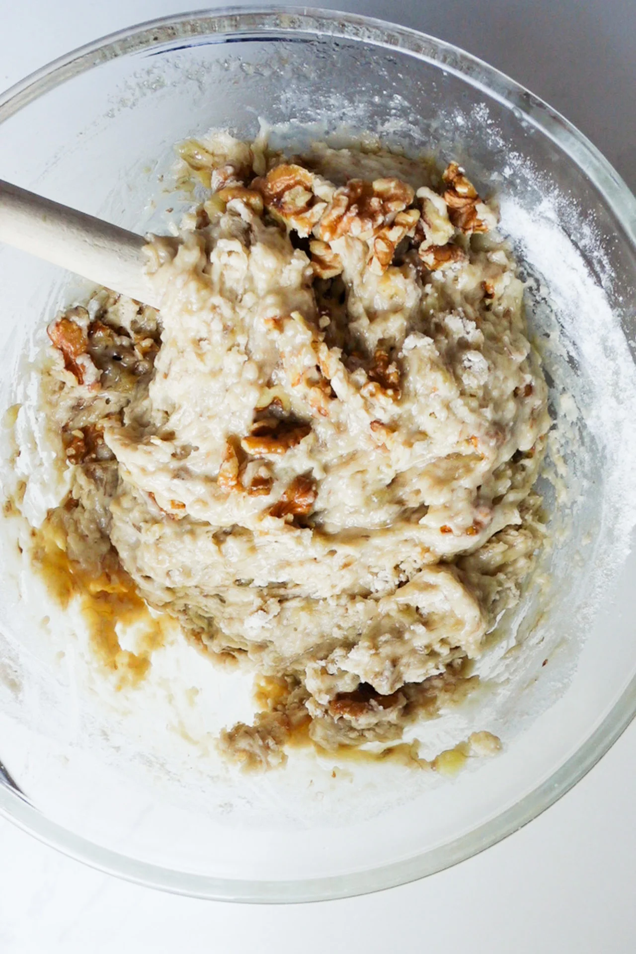 A glass bowl filled with moist banana bread batter, visible chunks of banana and walnuts, and some unmixed flour around the edges. A wooden spoon rests inside, showing the batter is being mixed by hand on a mostly clean white countertop.
