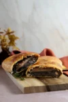 A golden-brown mushroom Wellington cut in half reveals a savory vegetarian Wellington filling inside flaky puff pastry. It sits on a wooden board with thyme, while a rust-colored cloth and blurred decorative plant are visible in the background.