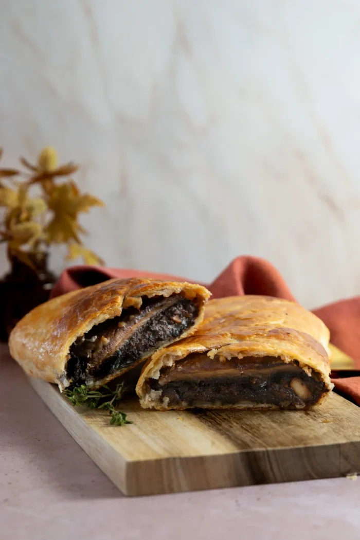 A golden-brown mushroom Wellington cut in half reveals a savory vegetarian Wellington filling inside flaky puff pastry. It sits on a wooden board with thyme, while a rust-colored cloth and blurred decorative plant are visible in the background.