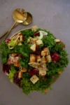 A bowl of mixed leafy greens, including kale and red lettuce, is topped with golden-brown garlic croutons. This Winter Green Salad sits on a light surface beside gold salad servers, the vibrant ingredients beautifully highlighted.