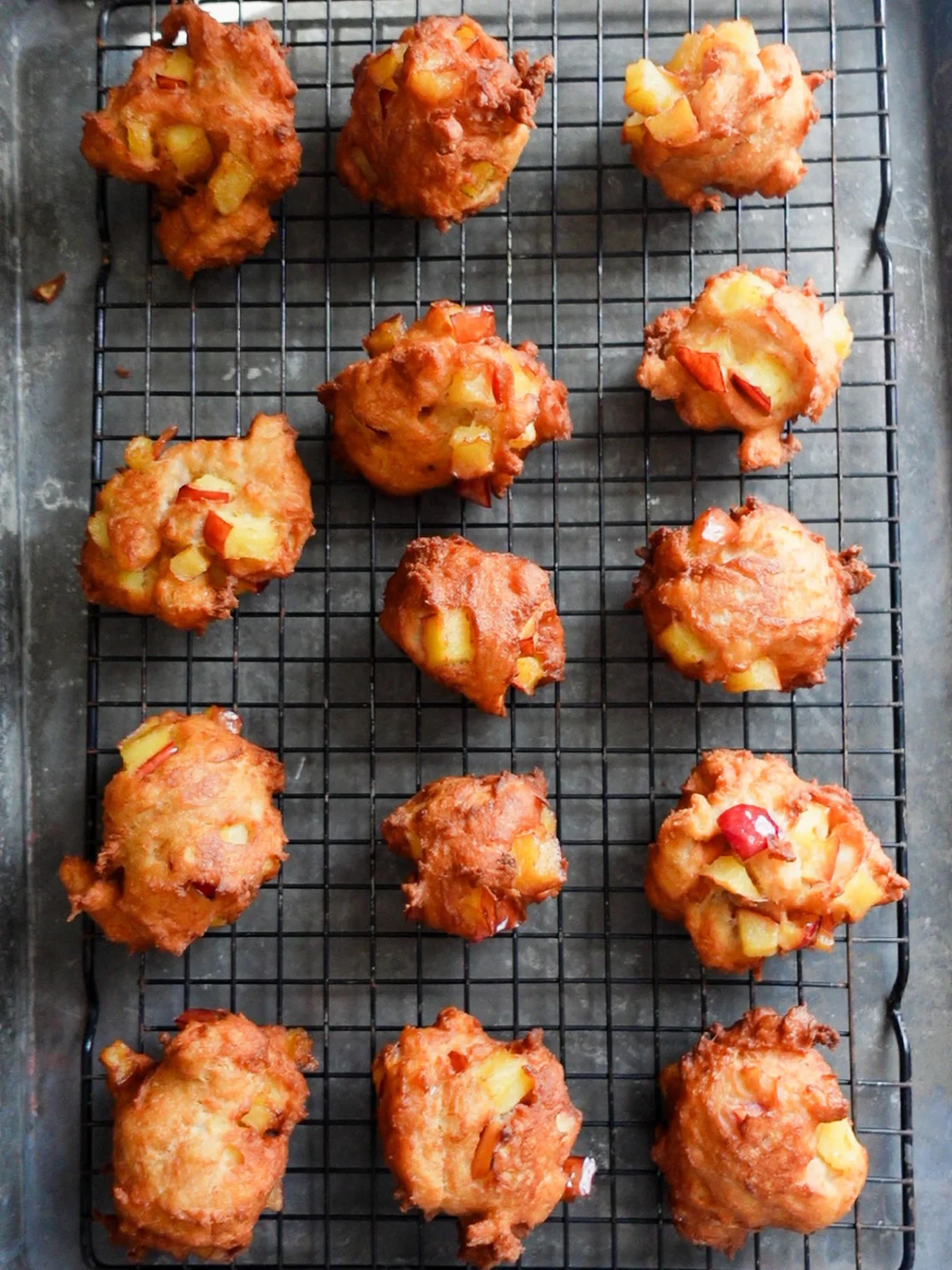 Twelve homemade apple fritters with crispy, golden-brown exteriors and visible chunks of apple rest on a black wire cooling rack over a dark baking tray. The unevenly shaped treats showcase red and yellow apple pieces throughout.