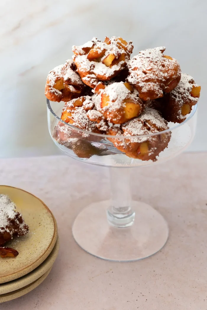 A glass pedestal dish holds a pile of crispy, golden-brown homemade apple fritters dusted with powdered sugar. Chunks of apple peek through the fritters. Nearby, stacked ceramic plates hold part of a fritter. The background is light and minimal.