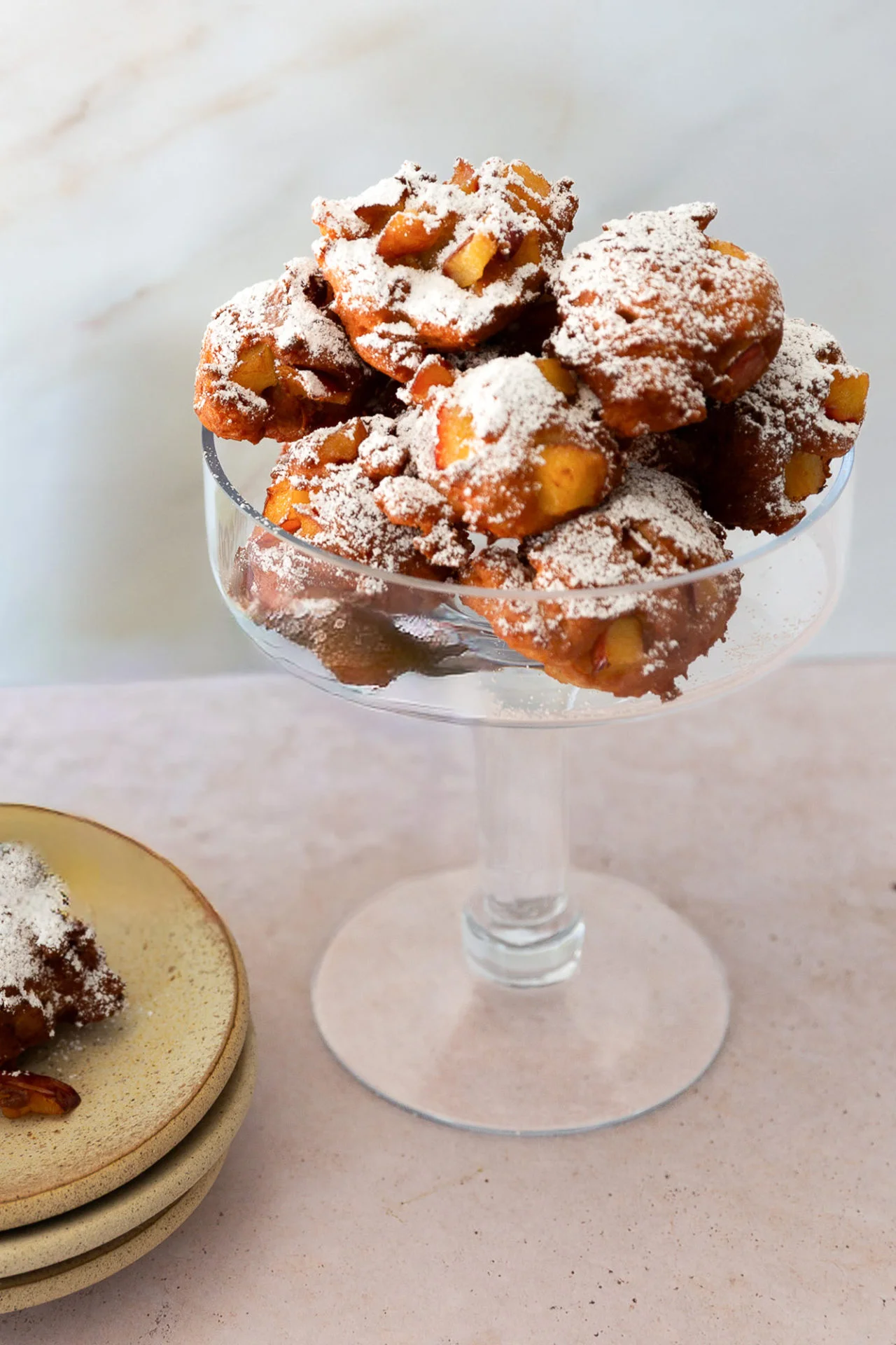 A glass pedestal dish holds a pile of crispy, golden-brown homemade apple fritters dusted with powdered sugar. Chunks of apple peek through the fritters. Nearby, stacked ceramic plates hold part of a fritter. The background is light and minimal.