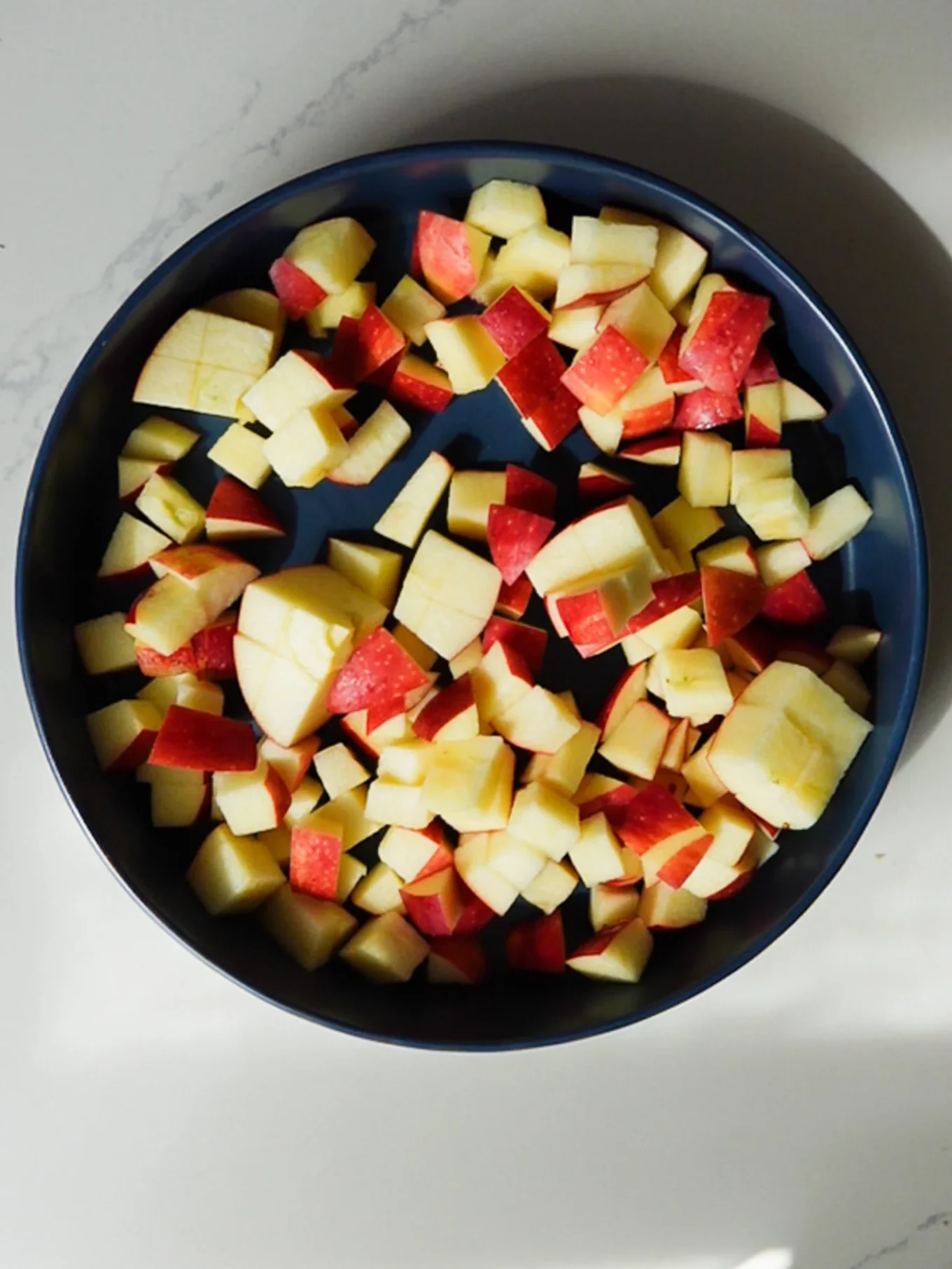 A round dark blue bowl filled with homemade, bite-sized cubes of red-skinned apples with pale yellow flesh sits on a light-colored, slightly marbled surface. The apple pieces vary in size and shape, reminiscent of apple fritters in the making.