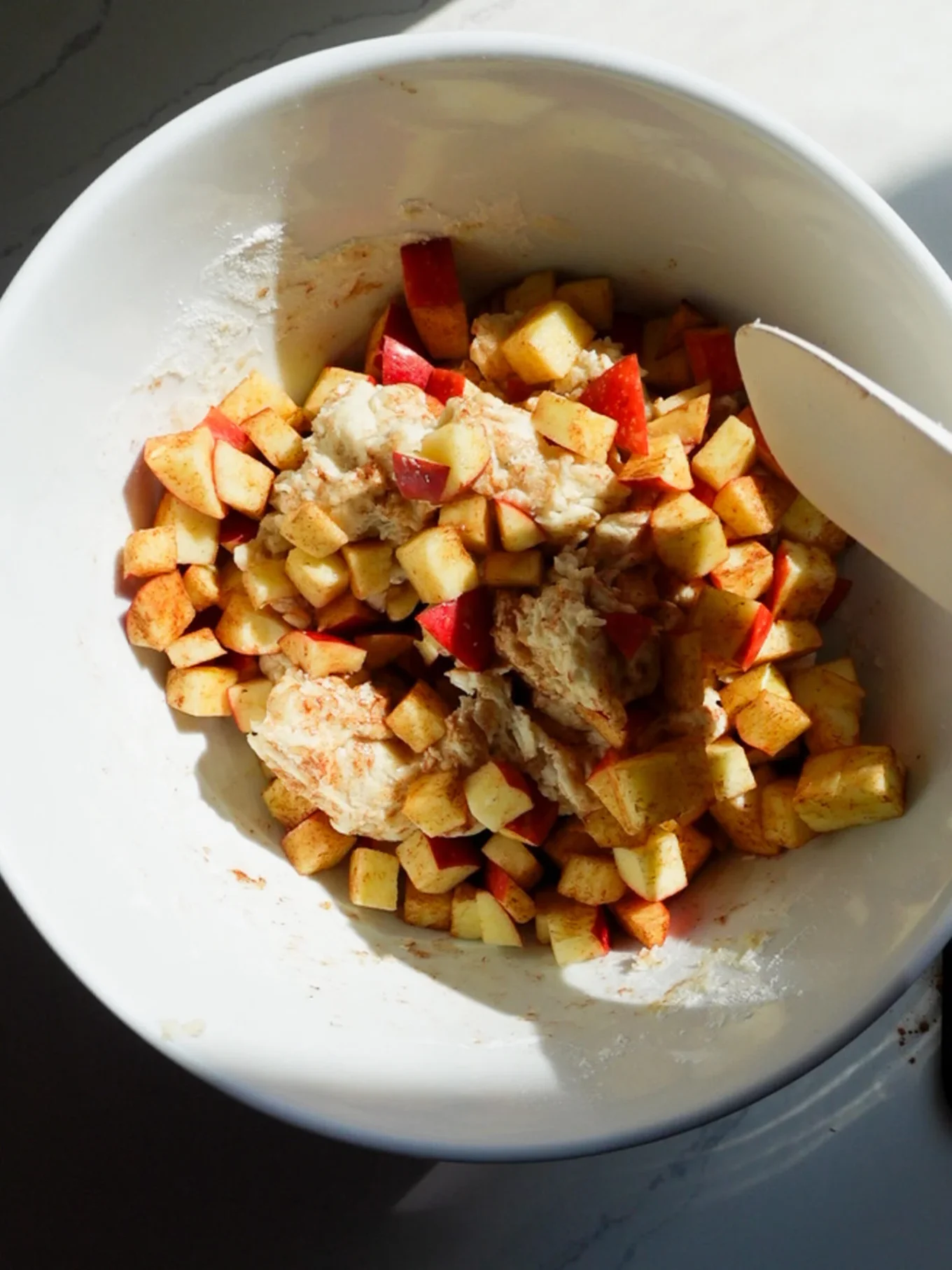 A white bowl filled with small, diced apple pieces mixed with cinnamon and dough—perfect for homemade Apple Fritters. A white spatula is partially visible on the right side, all on a white countertop in natural sunlight.