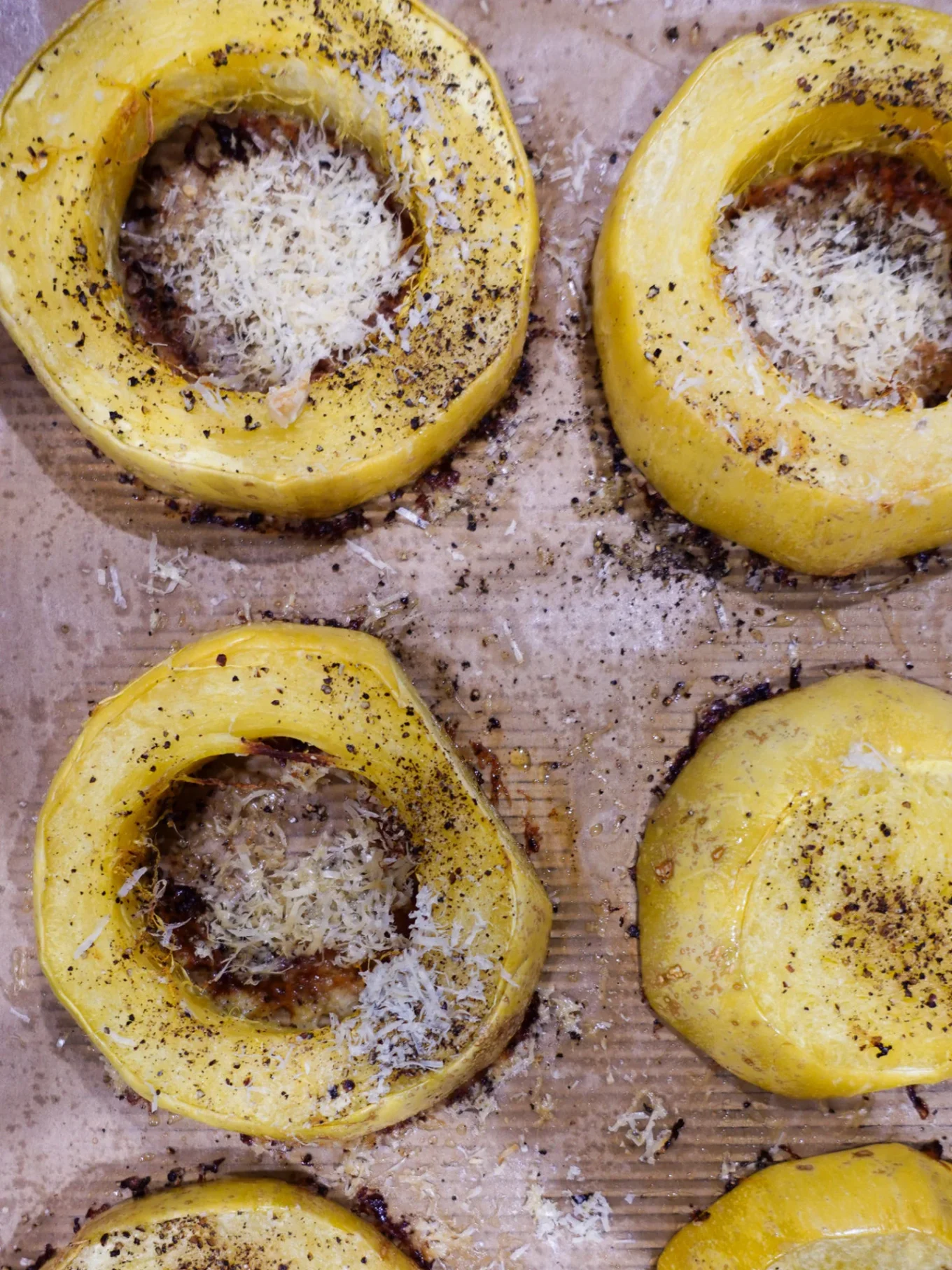 Six thick yellow squash rings are baked on a parchment-lined tray. Each ring’s center is filled with melted grated cheese, sprinkled with black pepper and herbs for a cacio e pepe twist. The squash has a roasted, slightly browned appearance.