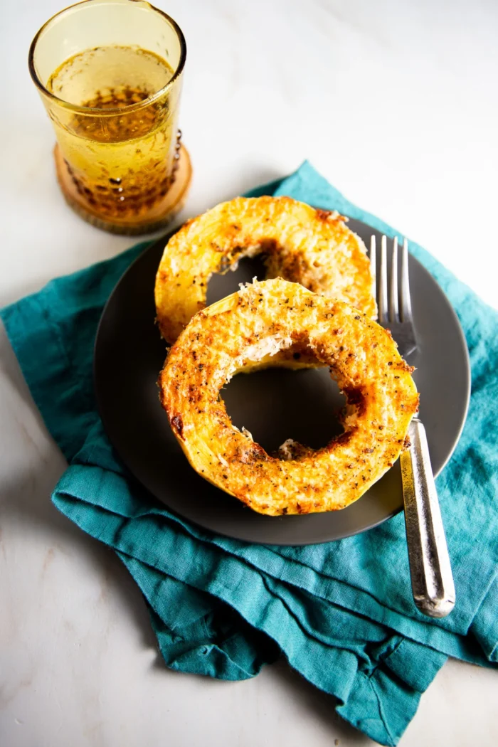 Two roasted spaghetti squash rings, inspired by Cacio e Pepe and seasoned with herbs and cheese, sit on a dark plate with a fork atop a turquoise cloth napkin. A tumbler of golden liquid rests on a coaster at the upper left.