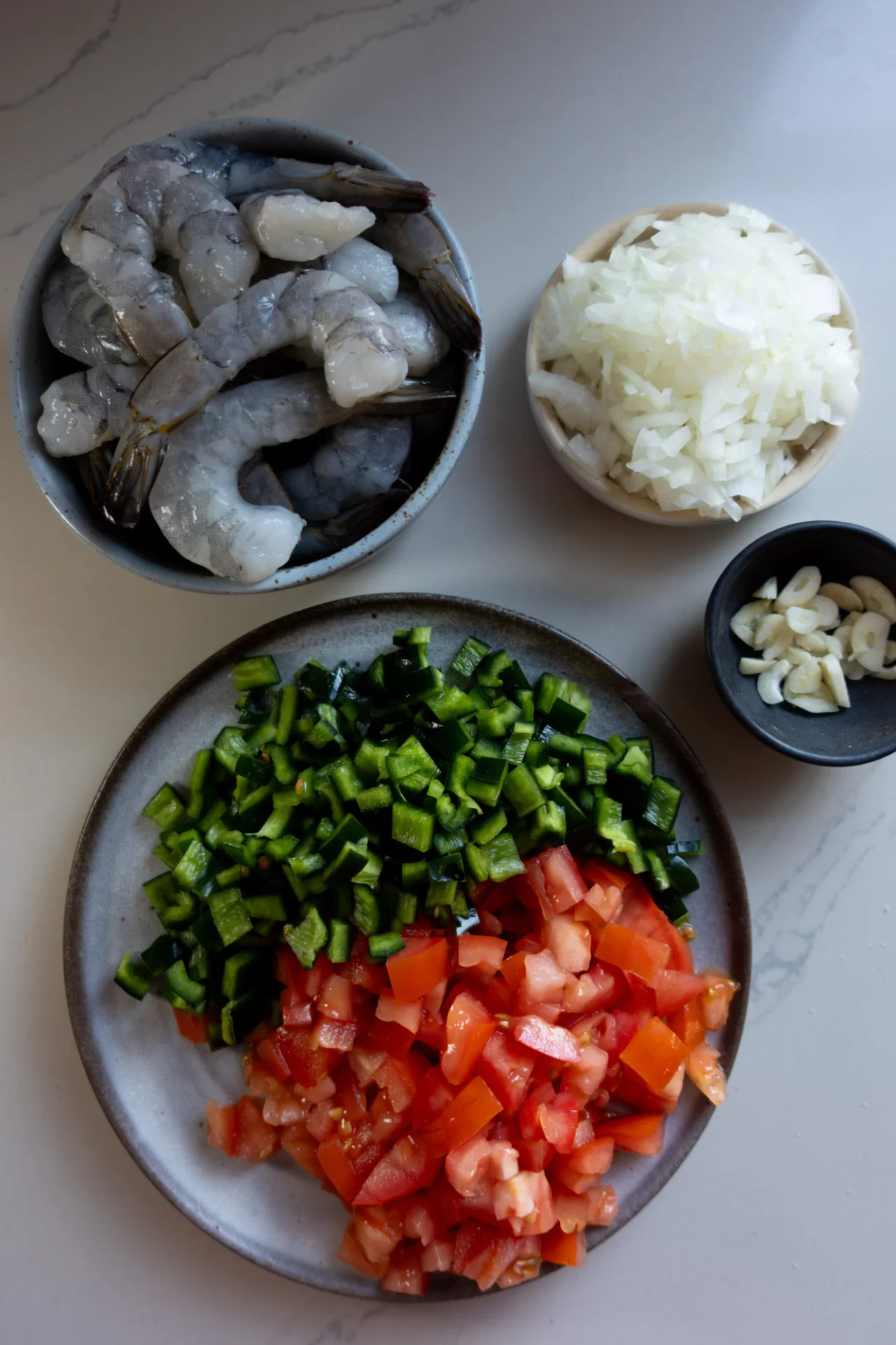 Four bowls and plates on a white surface display fresh ingredients for Shrimp Tacos: raw shrimp, chopped white onions, sliced garlic, and a plate with diced green bell peppers and tomatoes, perfect for making flavorful Mexican Tacos.