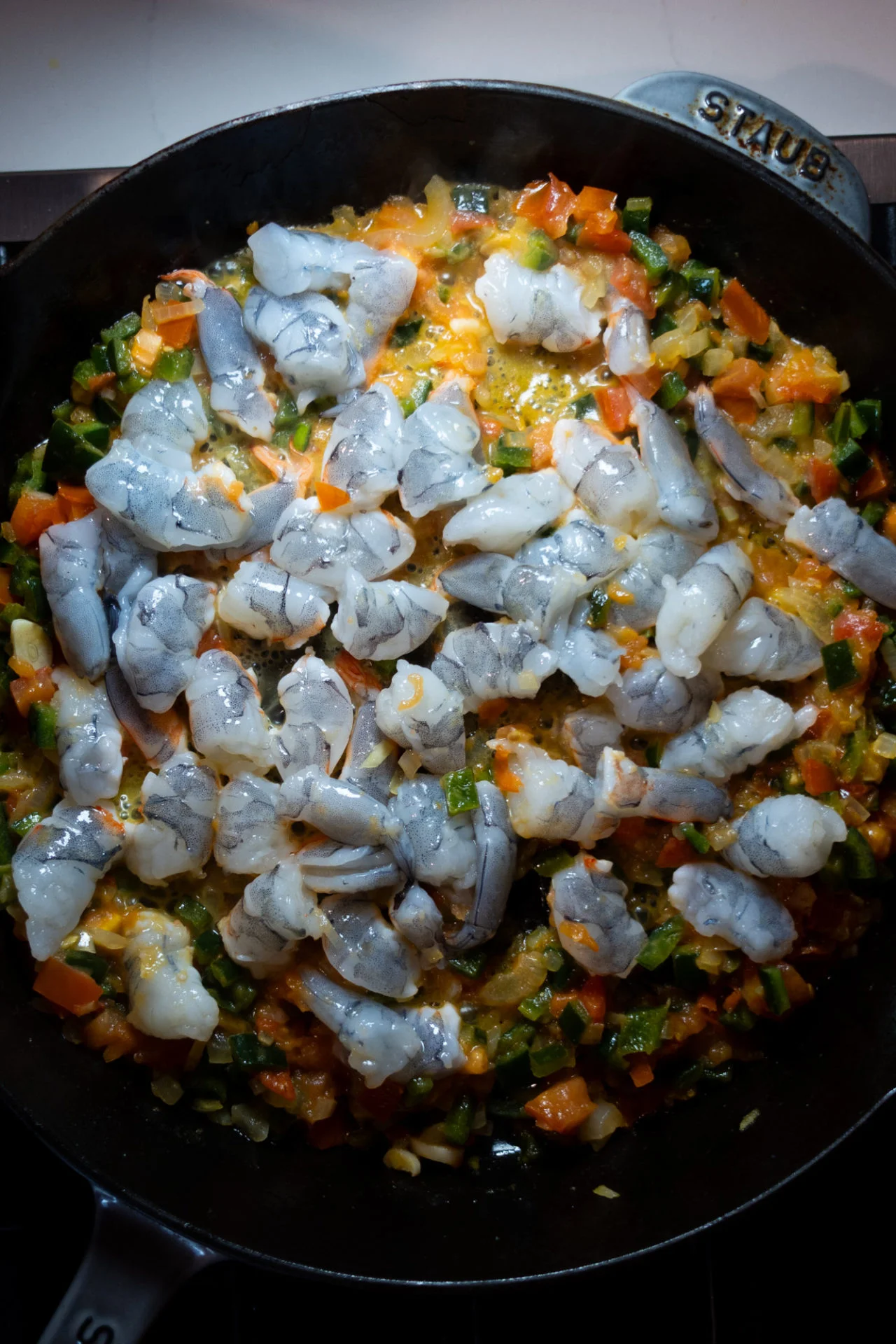 Raw shrimp are spread evenly in a cast iron skillet over simmering diced carrots, green bell peppers, onions, and yellow sauce—creating a vibrant mix perfect for Shrimp Tacos or inspired by classic Tacos Gobernador.