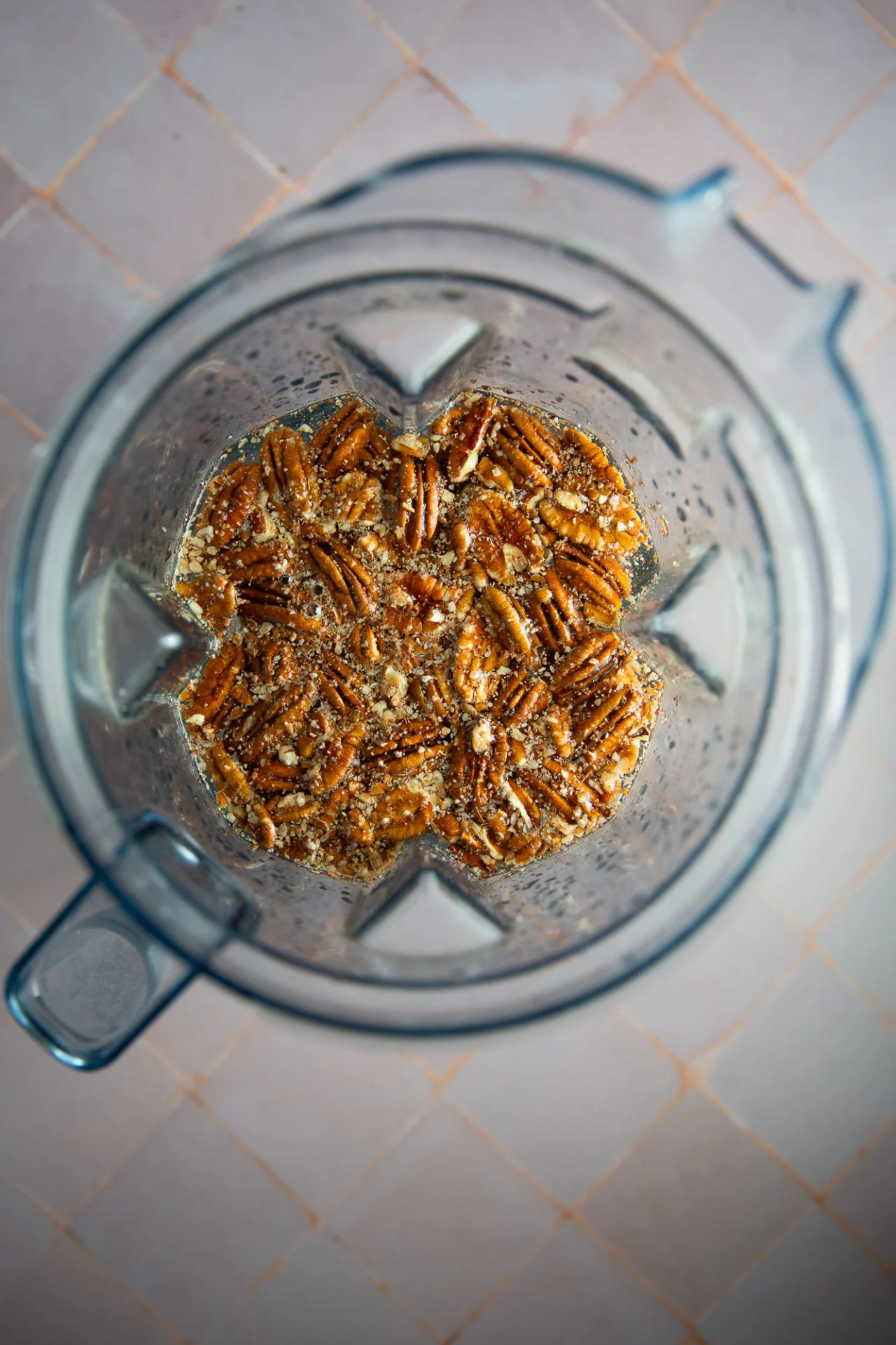 Overhead view of a blender containing pecans, chia seeds, and a liquid mixture, ready to be blended into creamy vegan pecan milk. The blender sits on a pinkish tiled surface with the ingredients clearly visible and unblended.