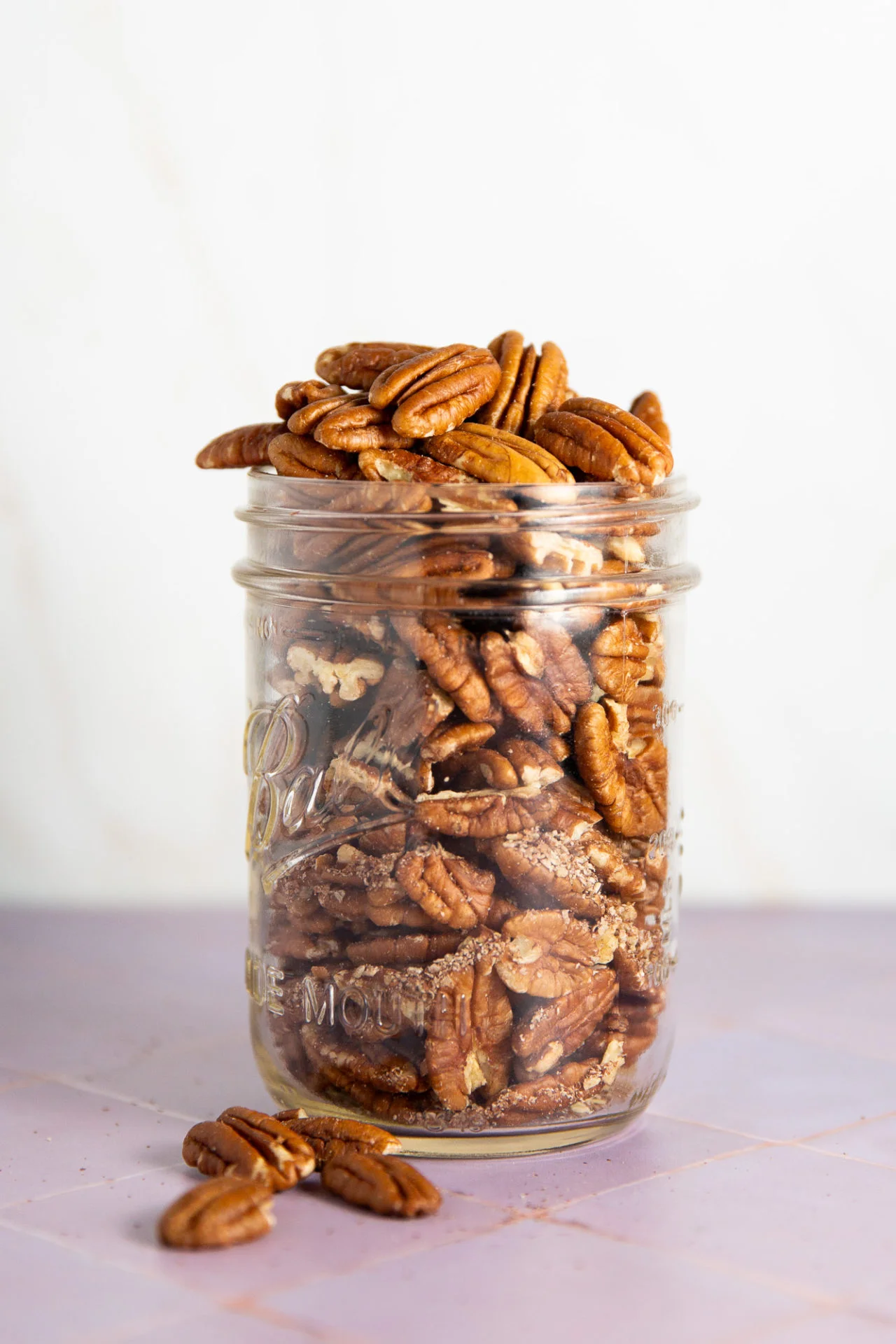 A clear glass jar filled to the top with pecan halves sits on a light purple surface. A few pecans are scattered in front, perfect for making creamy vegan pecan milk. The plain white background highlights their rich brown color and texture.